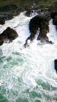 Vertical drone capture of strong waves breaking forcefully against steep rock formations at Sawarna Beach, Indonesia. Intense ocean movement and white sea foam create a dramatic tropical scene.