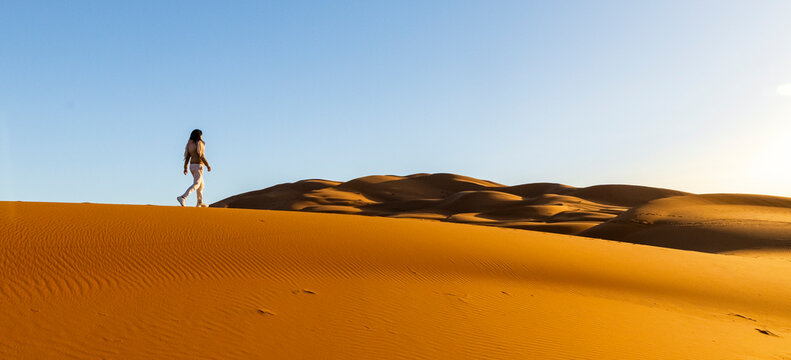 A woman walks across a large sand dune under a clear blue sky. Mezouga,Sahara desert,Morocco