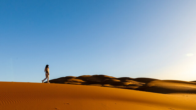 A woman walks along the crest of a large sand dune under a clear blue sky. Mezouga,Sahara desert,Morocco