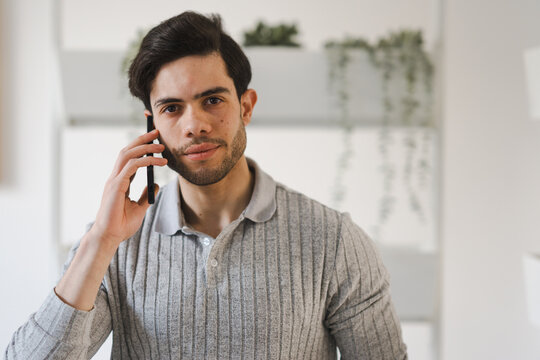 A young man with dark hair and a beard is talking on his mobile phone. He is wearing a grey polo shirt and looking directly at the camera.
