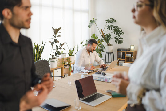 A man works on a laptop at a table while two colleagues converse in the foreground, suggesting a collaborative office environment.