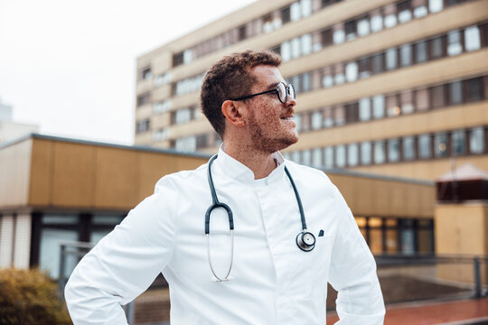 Doctor with stethoscope outdoors by hospital building looking away