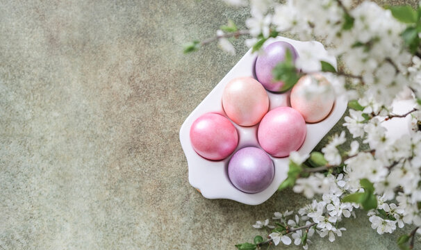 Top-down view of pastel pink and purple Easter eggs in white tray, framed by delicate white spring blossoms on textured background with copy space