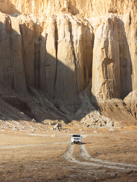 View of a lone white vehicle traversing a parched, cracked landscape towards towering, striated cliffs under the harsh sun, Shetpe, Mangystau Region, Kazakhstan.