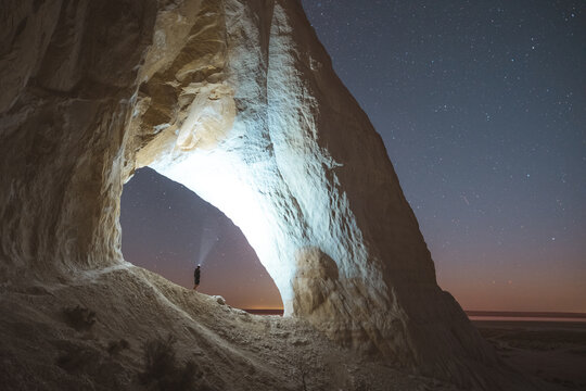 View of a person standing in a rock cave illuminated by a bright light under a starry night sky, Aktau, Mangystau Region, Kazakhstan.