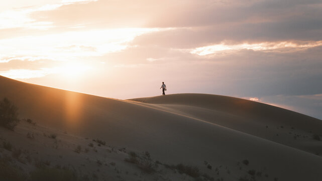 View of a lone figure atop a vast, undulating sand dune bathed in the warm glow of a setting sun, creating a serene desert landscape, Senek, Mangystau Region, Kazakhstan.