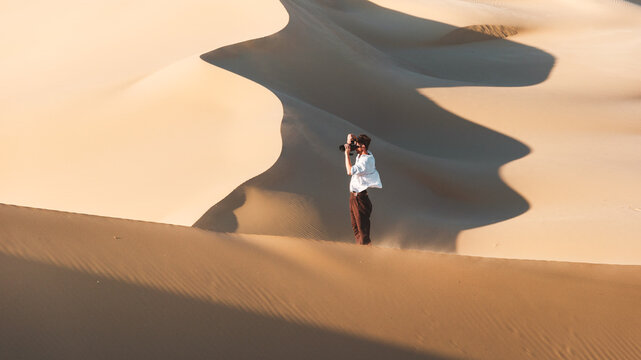 View of a lone figure capturing the undulating dunes, bathed in golden sunlight, as sand swirls around them, Senek, Mangystau Region, Kazakhstan.