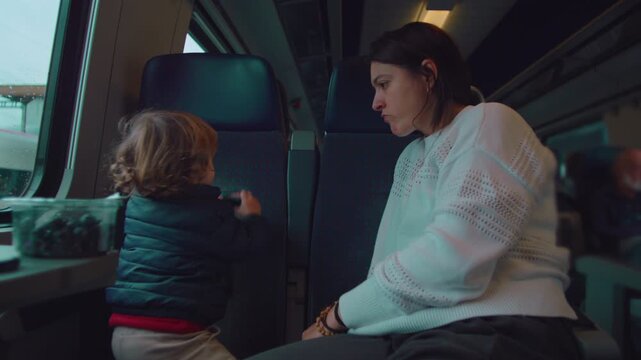 Toddler standing on train seat facing backrest playing with toy as mother watches attentively during commute conveying exploration guidance patience and everyday parenting routine