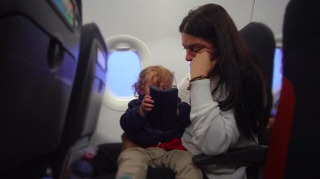 Fatigued mother seated on airplane holding toddler on lap while child explores object evoking patience caregiving strain and quiet preflight family travel moment