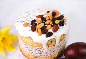 Traditional Easter cake with white icing, nut and raisin topping on light background. Homemade festive pastry with spring flowers and dyed egg. Cozy holiday food concept. Selective focus.