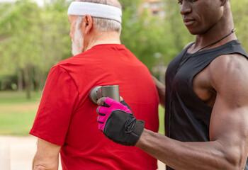 Naklejka premium Close up of Strong personal trainer giving massage therapy with gun to athletic elderly man outdoors