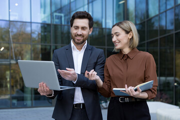 Two smiling business professionals engaging in a discussion while looking at a laptop, standing together outside a modern office building, symbolizing teamwork and successful partnership