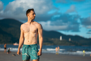 handsome white man with athletic muscular body standing at beach at sunny sunset, attractive male athlete walking at sea or ocean shore