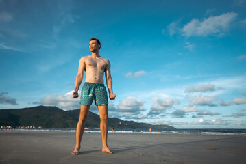 handsome white man with athletic muscular body standing at beach at sunny sunset, attractive male athlete walking at sea or ocean shore