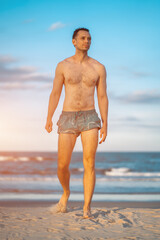 handsome white man with athletic muscular body standing at beach at sunny sunset, attractive male athlete walking at sea or ocean shore