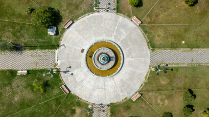 Symmetrical zenithal shot of a circular fountain set within a park. The central water feature is surrounded by concentric rings of concrete and green lawn areas intersected by geometric paved paths. © Stefano Tammaro