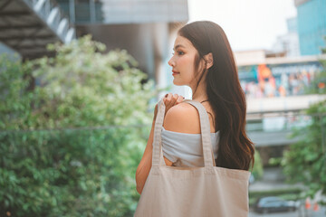 Beautiful Asian woman carrying reusable canvas tote bag while walking in the city, eco-friendly lifestyle and plastic-free concept, young stylish lady with fabric bag shopping outdoors, sustainable