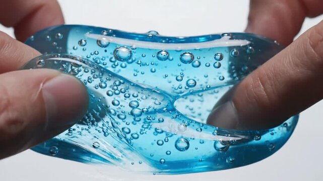Close-up, slow-motion video of hands gently playing with a translucent blue gel containing air bubbles, highlighting its elasticity.