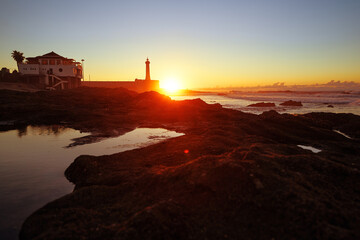 Beautiful twilight setting featuring lighthouse in distance, radiant sunset glow over ocean waves, and rocky shoreline foreground with reflections creating a serene and picturesque coastal atmosphere.