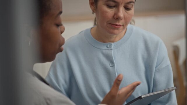 Diverse female doctor and patient discussing medical test results on digital tablet during professional appointment in modern clinic setting