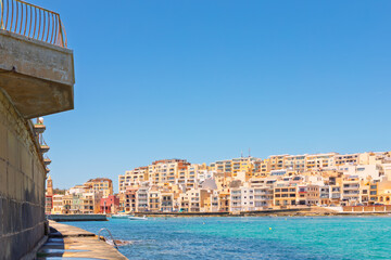 Waterfront of Marsaskala, Malta, featuring colorful residential buildings along the Mediterranean coast. A stone promenade and turquoise waters define this tranquil southern bay