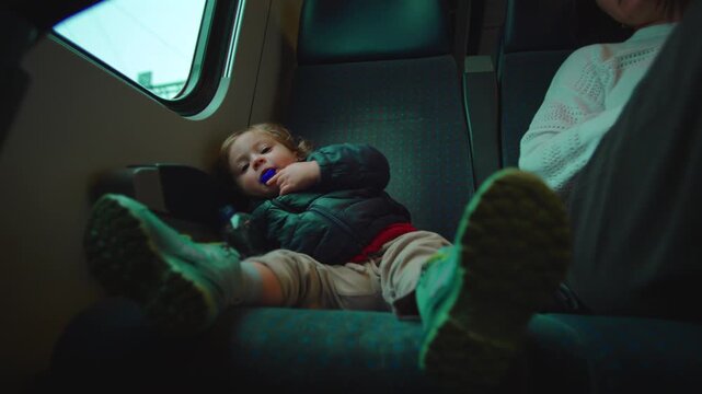 Toddler resisting parent during train ride, child chewing bottle cap on seat, playful defiance and concern mix, candid family travel moment showing curiosity, stubbornness, care