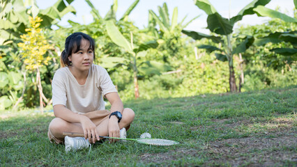 A girl sitting on the grass with a badminton racket, looking serious in an outdoor garden.