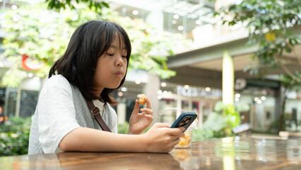 A girl eating snacks while looking at her smartphone in an outdoor mall.