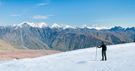 Hiker standing on snowy mountain slope looking at dramatic rugged peaks under clear blue sky.