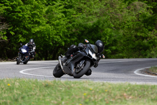 Motorcyclist in black gear leaning into a sharp corner on a scenic winding road through a lush green forest.