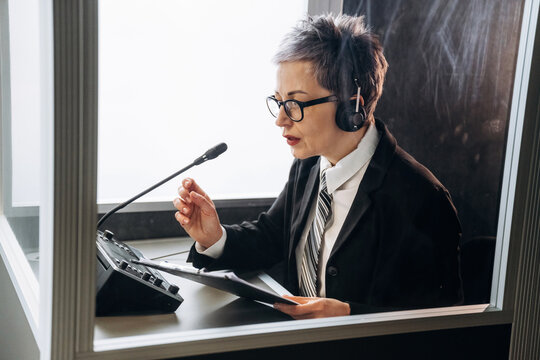 Female interpreter wearing headset speaks into microphone while seated at desk with audio equipment in a soundproof booth during a conference setting