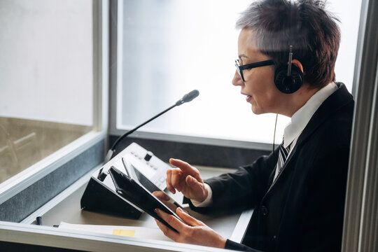 Female interpreter wearing headset and suit uses tablet while working in soundproof booth during conference, microphone and control panel visible in the background