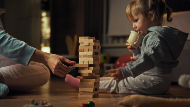 Young daughter and her mother are immersed in playful learning on the floor, constructing precarious tower using wooden Jenga blocks, enjoying interactive evening of balance and fun together in warm