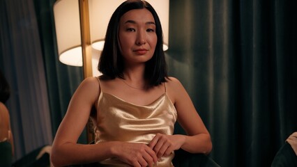 Fototapeta premium Portrait calm serious Asian woman sitting at home evening close up Japanese Korean Chinese female indoors looking at camera confident wearing golden dress ready for party date look gaze beauty style
