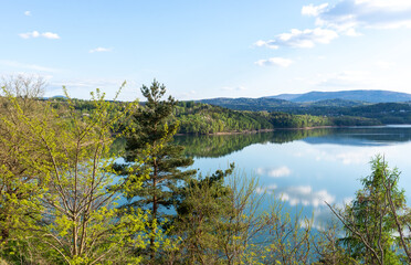 Wide view of lake dobczyckie with pine tree and mixed forest reflected in still water. Rolling hill and distant mountain create layered composition under bright sky, highlighting serene outdoor scener