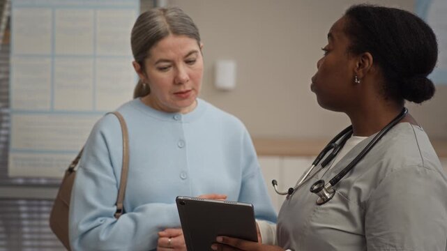 Diverse female patient and doctor having detailed discussion while viewing health test results on digital tablet in professional medical setting