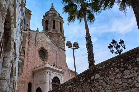 Historic church facade with bell tower and palm tree rising above stone staircase in old town sitges under dramatic cloudy sky.