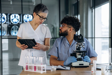 Fototapeta premium Female healthcare professional in white uniform discusses findings with male colleague in blue scrubs at laboratory table with microscope and test tubes in modern medical facility