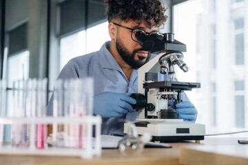 Male scientist wearing blue lab coat and gloves examines samples under a microscope in a modern...