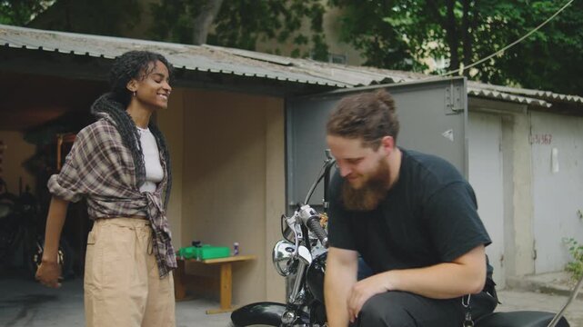 A young couple stands near a garage, playing rock paper scissors to decide who will ride the motorcycle. The woman wins, creating a playful and lighthearted moment
