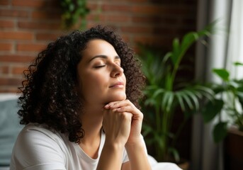 A young woman with curly dark hair is meditating with her eyes closed and hands clasped in front of her face, finding peace and serenity in a calm indoor setting