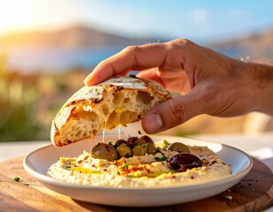 Hand Dipping Crusty Bread Into Creamy Hummus With Olives Garnished With Spices Outdoors With Scenic Sea View In Sunlight