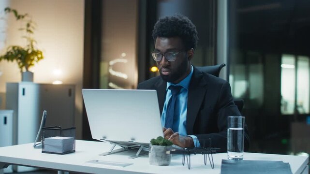 Businessman working late typing on laptop finishing tasks at office desk in evening wearing business suit focused expression managing workload indoors meeting deadlines updating reports efficiently.