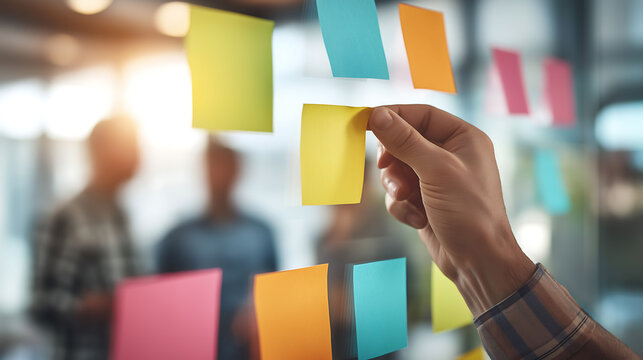 A hand places a colorful sticky note on a glass board during a team meeting, representing brainstorming, collaboration, and creative project planning in a modern office
