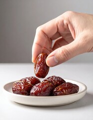 Hand selecting a single dark brown Medjool date from a white plate full of dates against a neutral gray background studio lighting