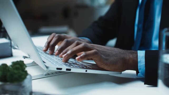Close up male hands typing on laptop keyboard while working late at office desk with business documents nearby. Focused professional finishing tasks on computer in modern workspace during evening.