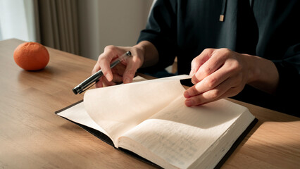 ノートに書き物をする男性の手元 思考の整理 ジャーナリング 丁寧な暮らし / Hands writing in notebook with pen and orange on table, journaling and planning concept
