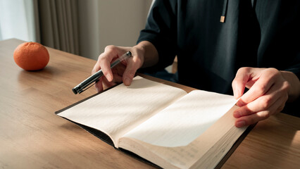 ノートに書き物をする男性の手元 思考の整理 ジャーナリング 丁寧な暮らし / Hands writing in notebook with pen and orange on table, journaling and planning concept