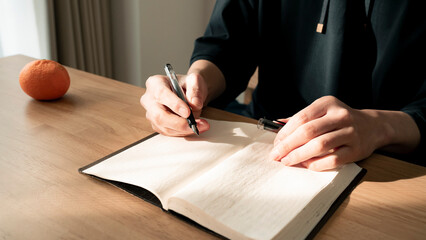 ノートに書き物をする男性の手元 思考の整理 ジャーナリング 丁寧な暮らし / Hands writing in notebook with pen and orange on table, journaling and planning concept