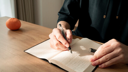 ノートに書き物をする男性の手元 思考の整理 ジャーナリング 丁寧な暮らし / Hands writing in notebook with pen and orange on table, journaling and planning concept